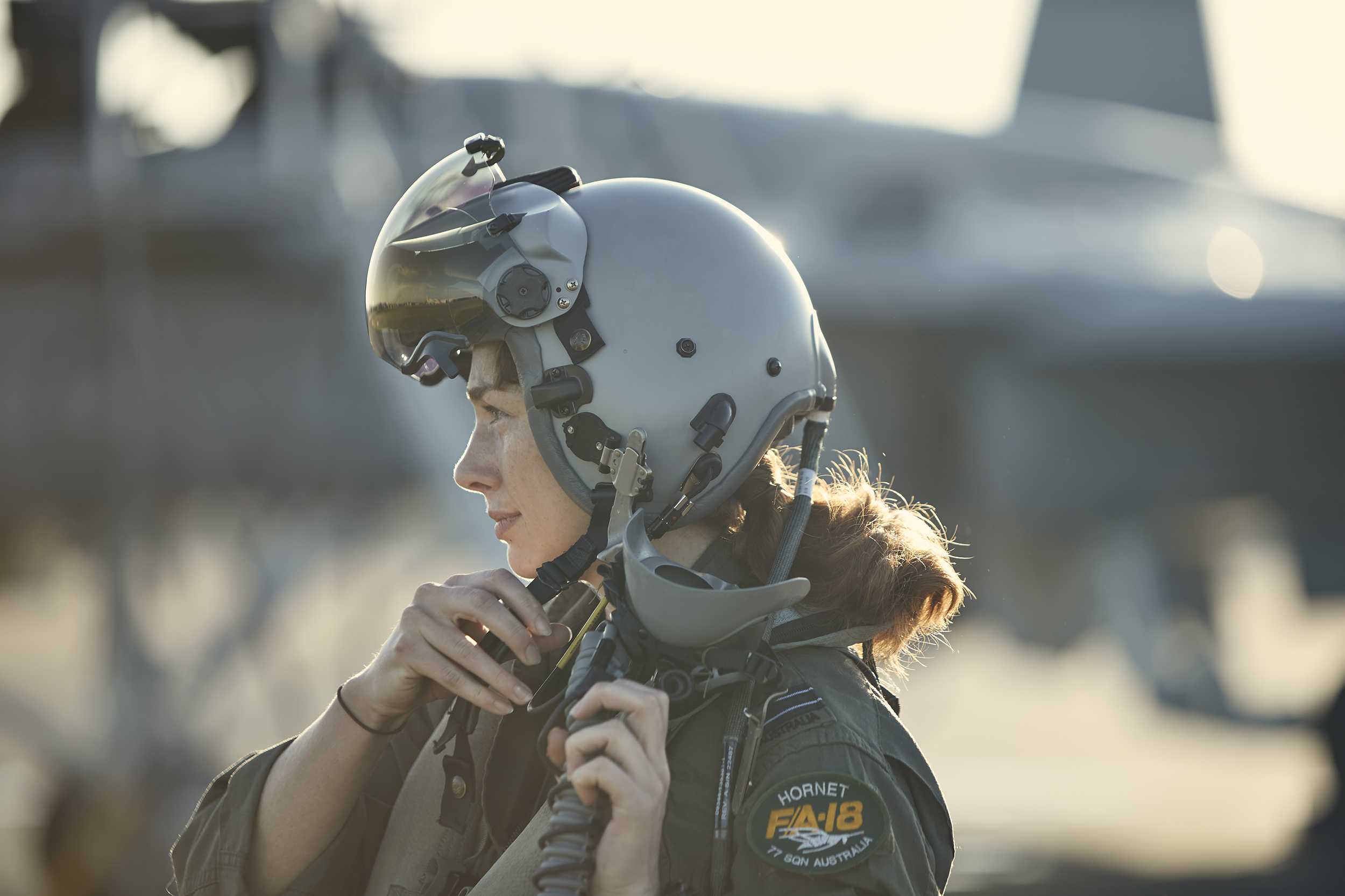 A fair-skinned woman in a pilot helmet and flight suit is the primary focus of this outdoor medium shot with a jet in the background.