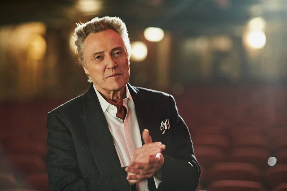 Portrait of a mature Caucasian man in a black jacket and white shirt, with hands clasped, set against a blurry theatre backdrop.