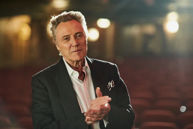 Portrait of a mature Caucasian man in a black jacket and white shirt, with hands clasped, set against a blurry theatre backdrop.