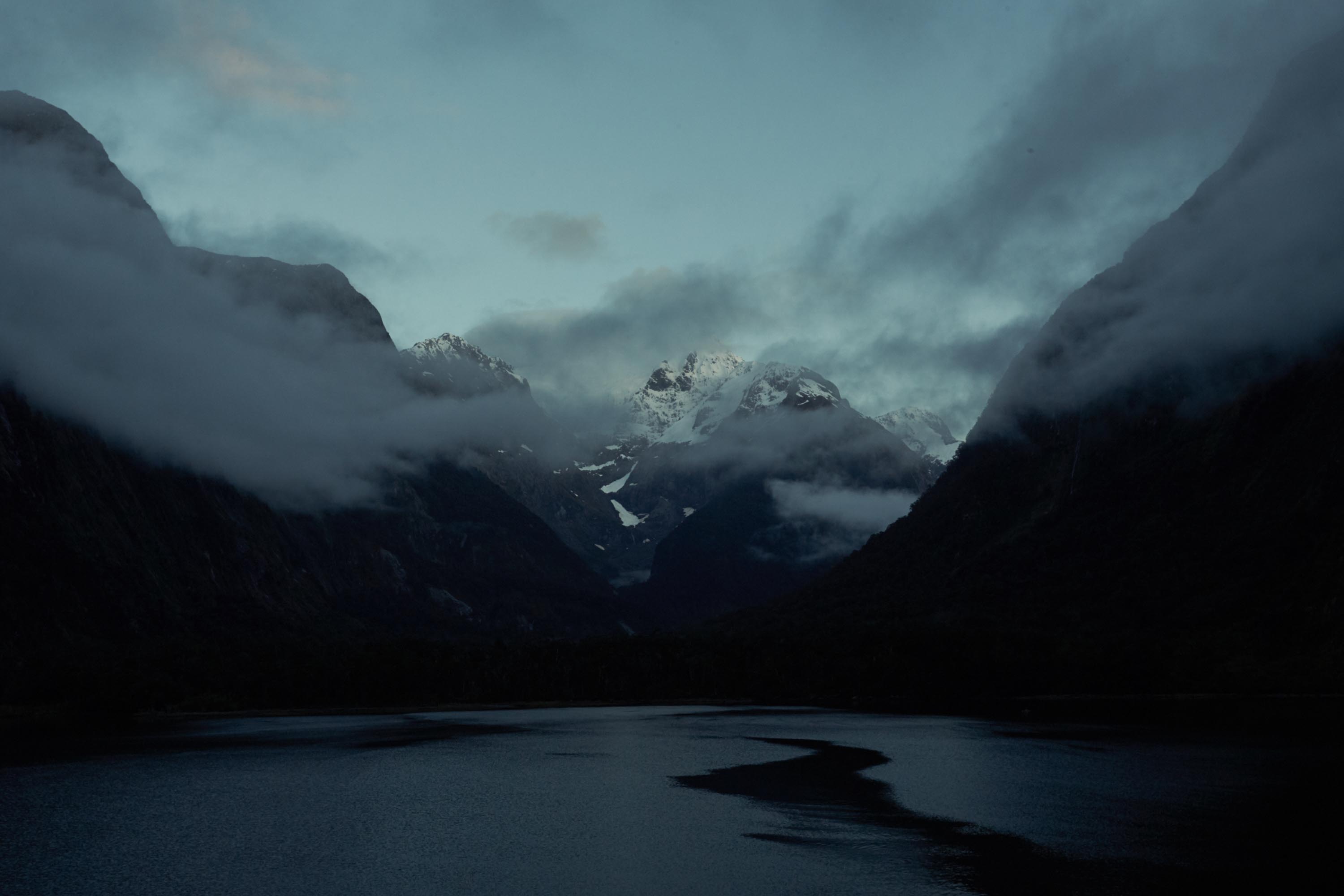 Tranquil, dark landscape view of snow-capped mountain peaks through the clouds, seen across a still, dark lake in shadow.