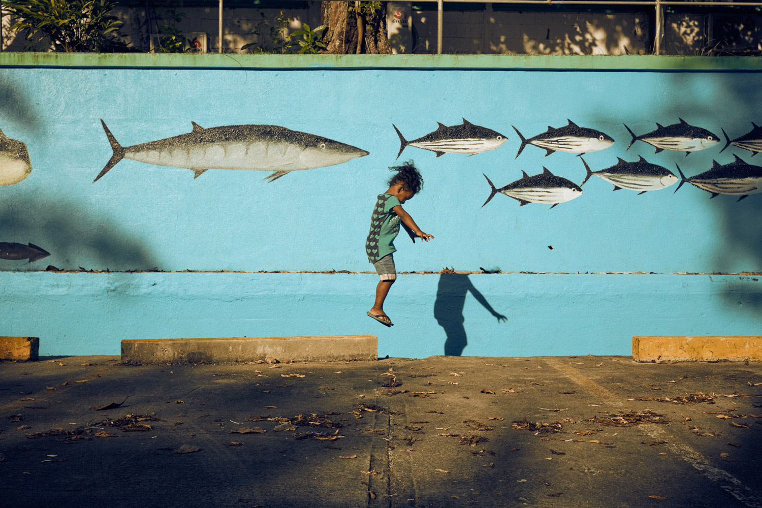 Child jumps in front of mural featuring fish on blue wall, creating a playful scene in an urban environment on a sunny day.