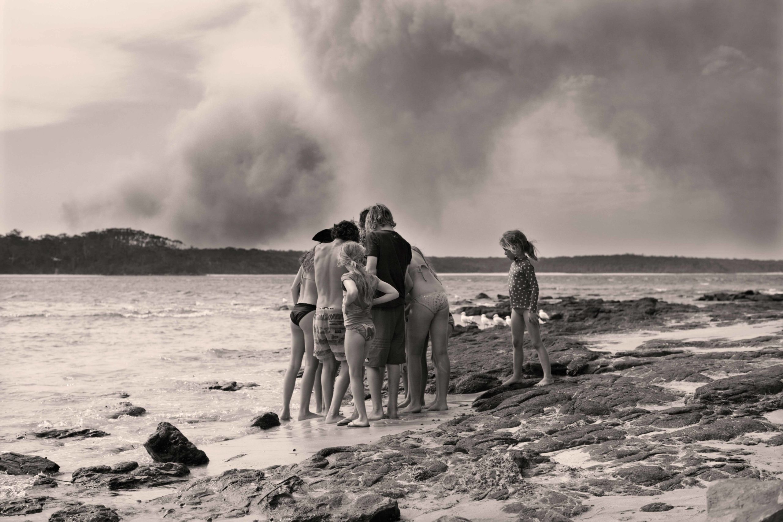 Group of adults and children standing on a rocky beach with ocean and cloudy sky background in a monochrome outdoor shot.