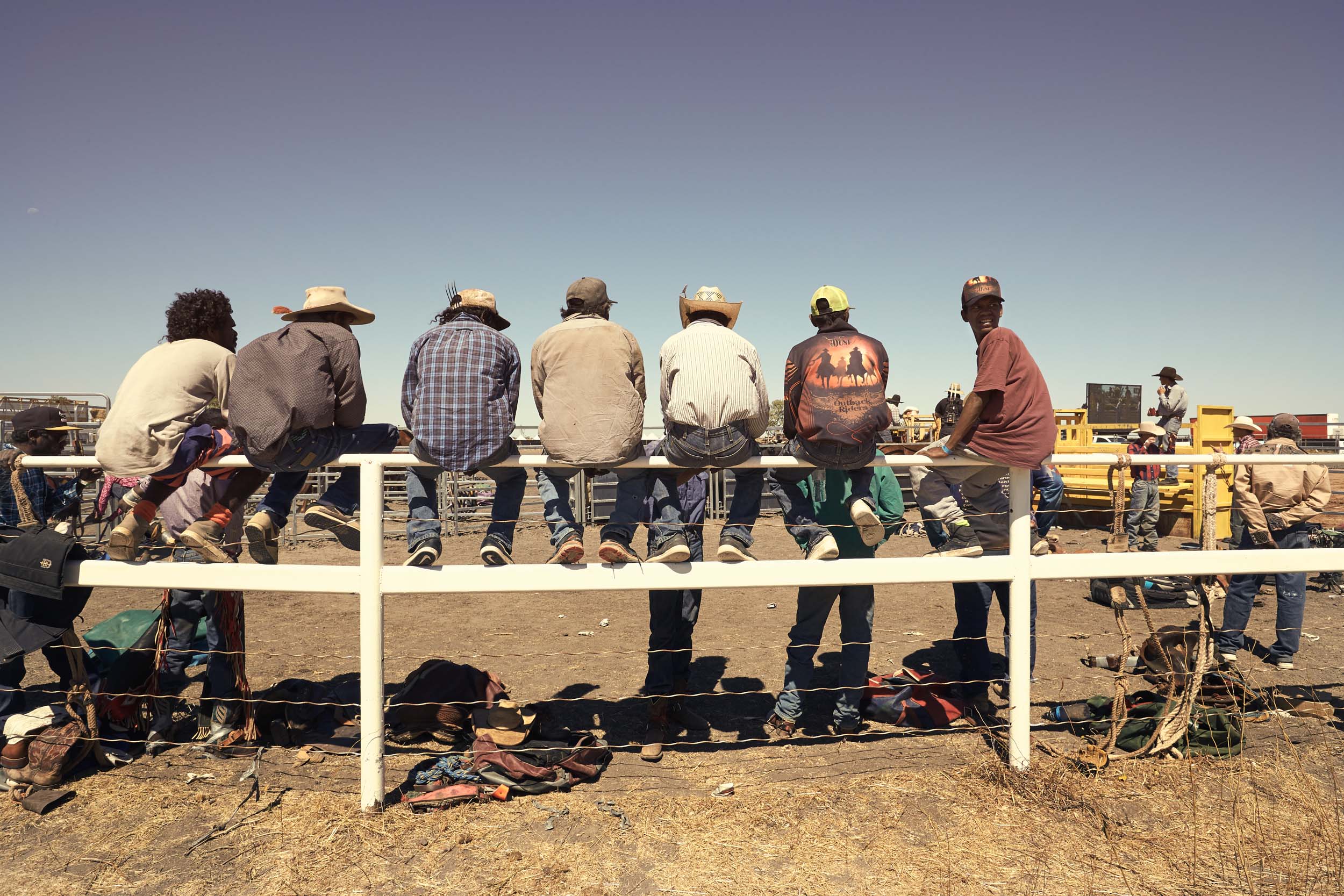 Group of people sitting on a white fence in an outdoor setting, possibly at a rodeo or rural event, under a clear blue sky.