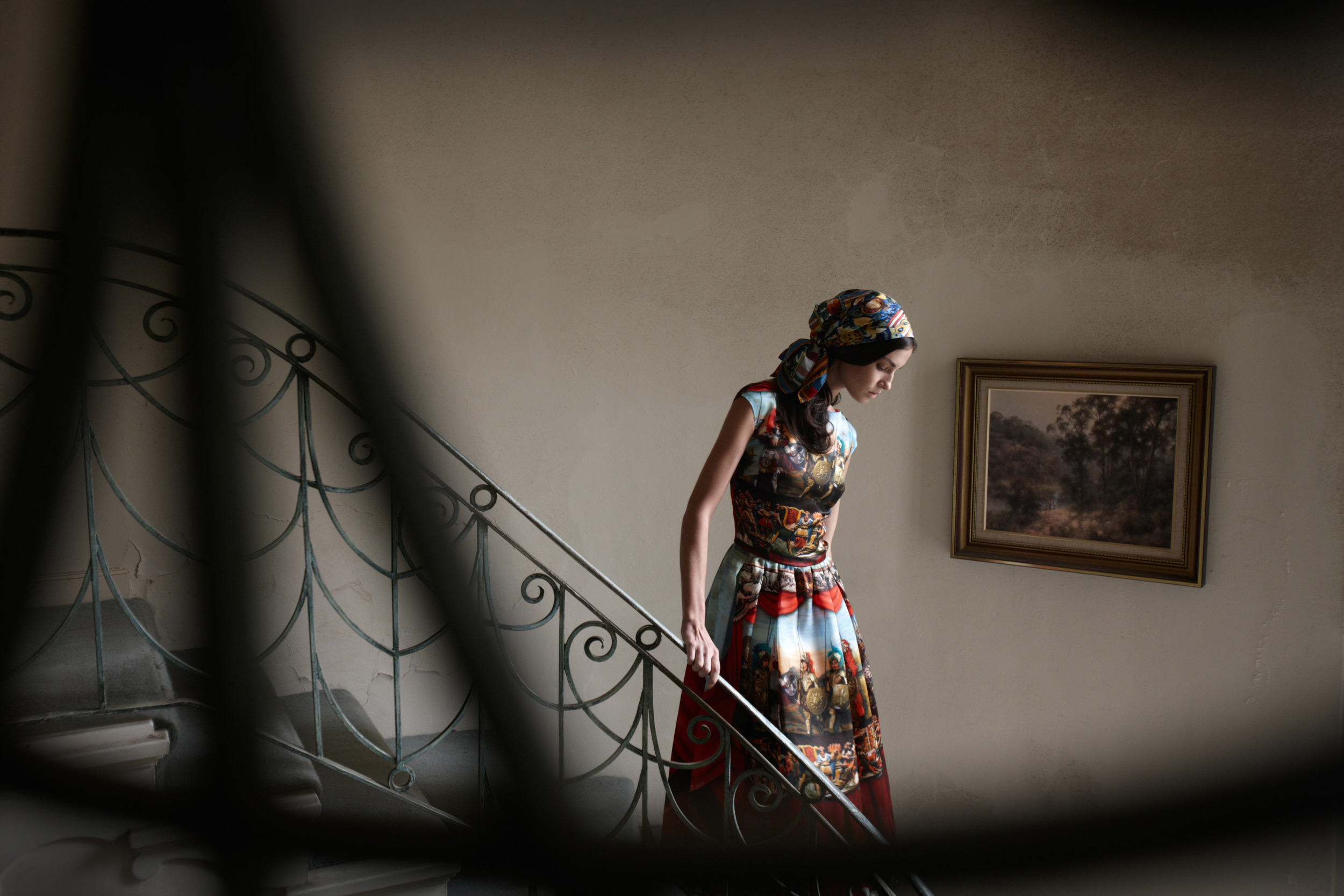 Woman in patterned dress and headscarf descends patterned staircase in indoor setting; framed landscape painting hangs on the wall.