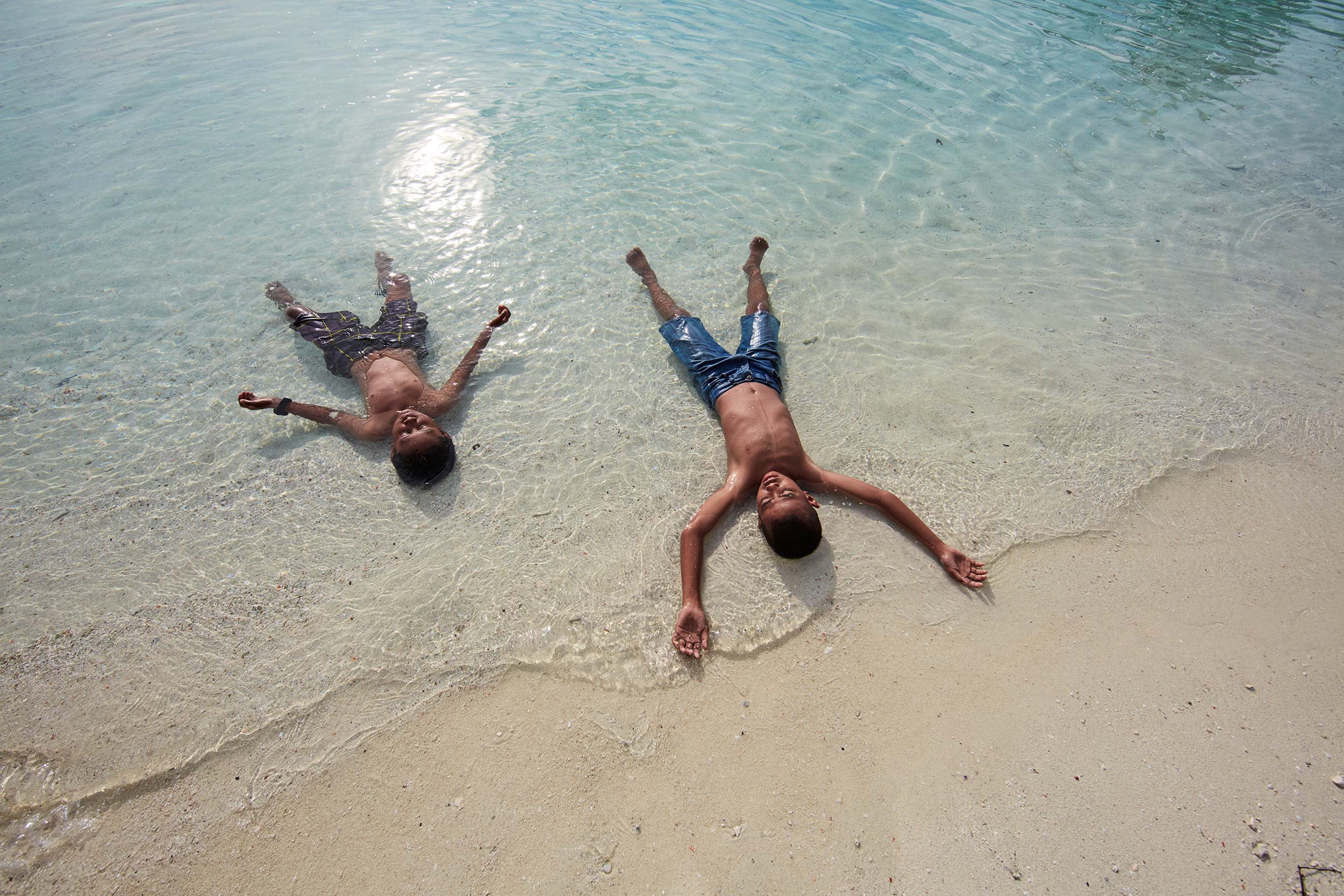 Two boys relaxing in shallow, clear water on a sunny day at the beach. The water meets the light-colored sand along the shoreline.