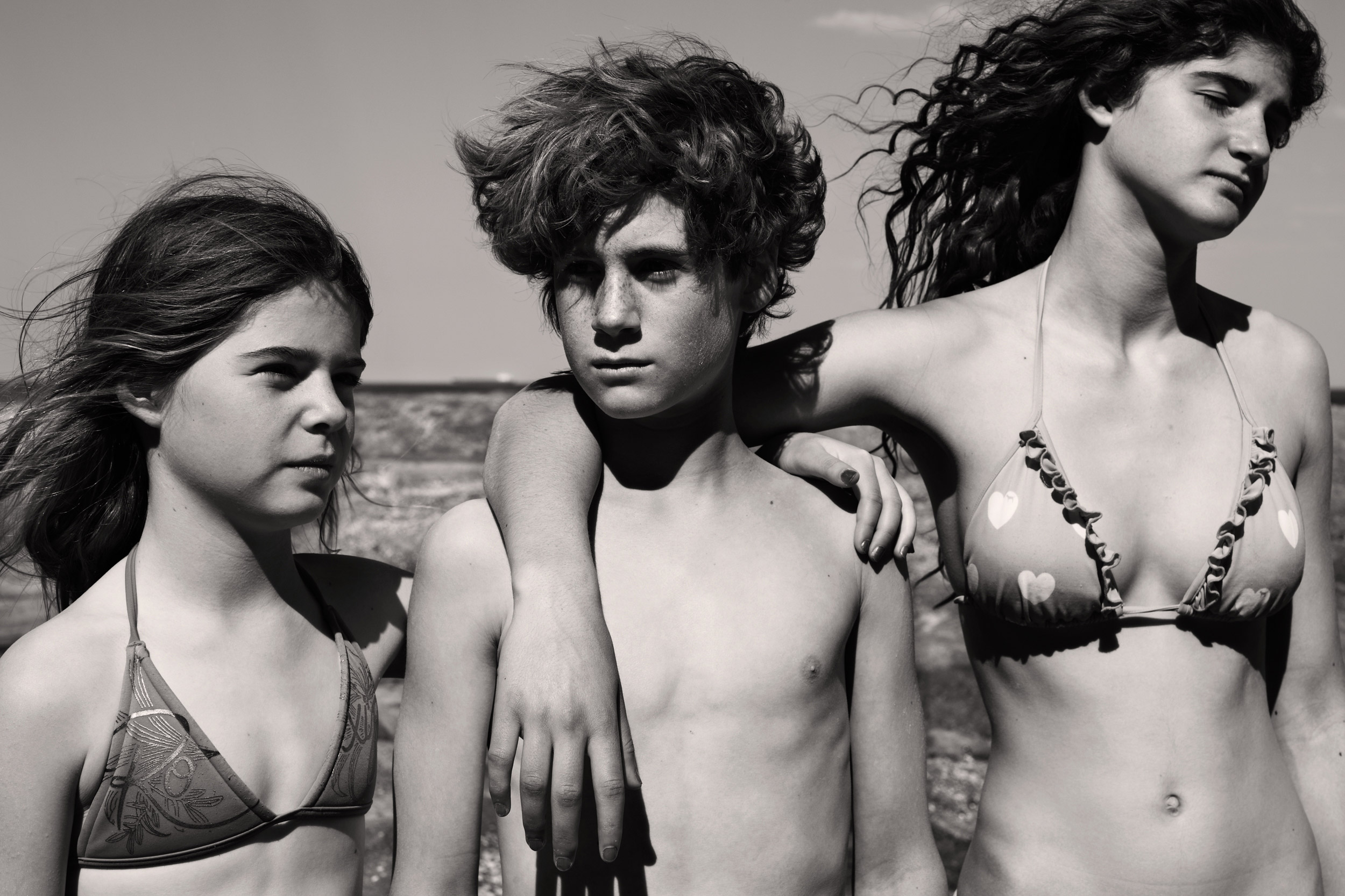 Black and white shot of three young people in swimsuits standing outside with a background of blue sky and neutral toned sand.