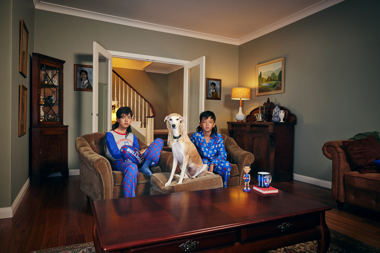 Two children and a dog sit on a couch in pajamas in a brightly lit living room with hardwood floors and brown furniture.