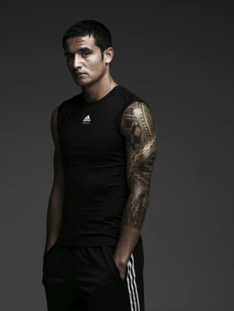 Man wearing a black sleeveless athletic top with extensive tribal arm tattoos standing against a grey background.