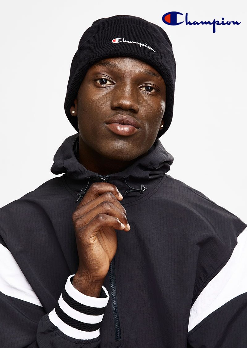 A dark-skinned adult male wearing a black Champion beanie and hooded jacket poses indoors against a white backdrop in a studio shot.