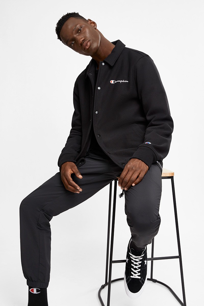 Man in black Champion jacket and black pants sitting on a stool in a white studio. He is wearing Converse sneakers and black socks.