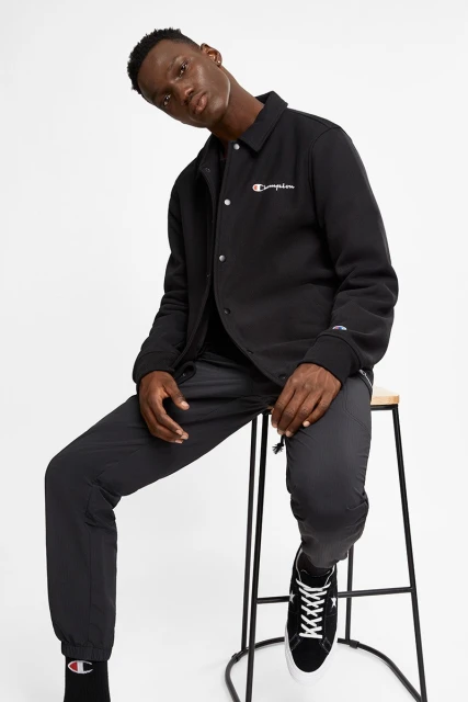 Man in black Champion jacket and black pants sitting on a stool in a white studio. He is wearing Converse sneakers and black socks.