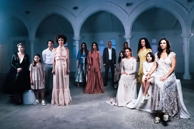 Group portrait of eleven women, two children, and one man in a room with arched doorways and pale walls, creating a formal composition.