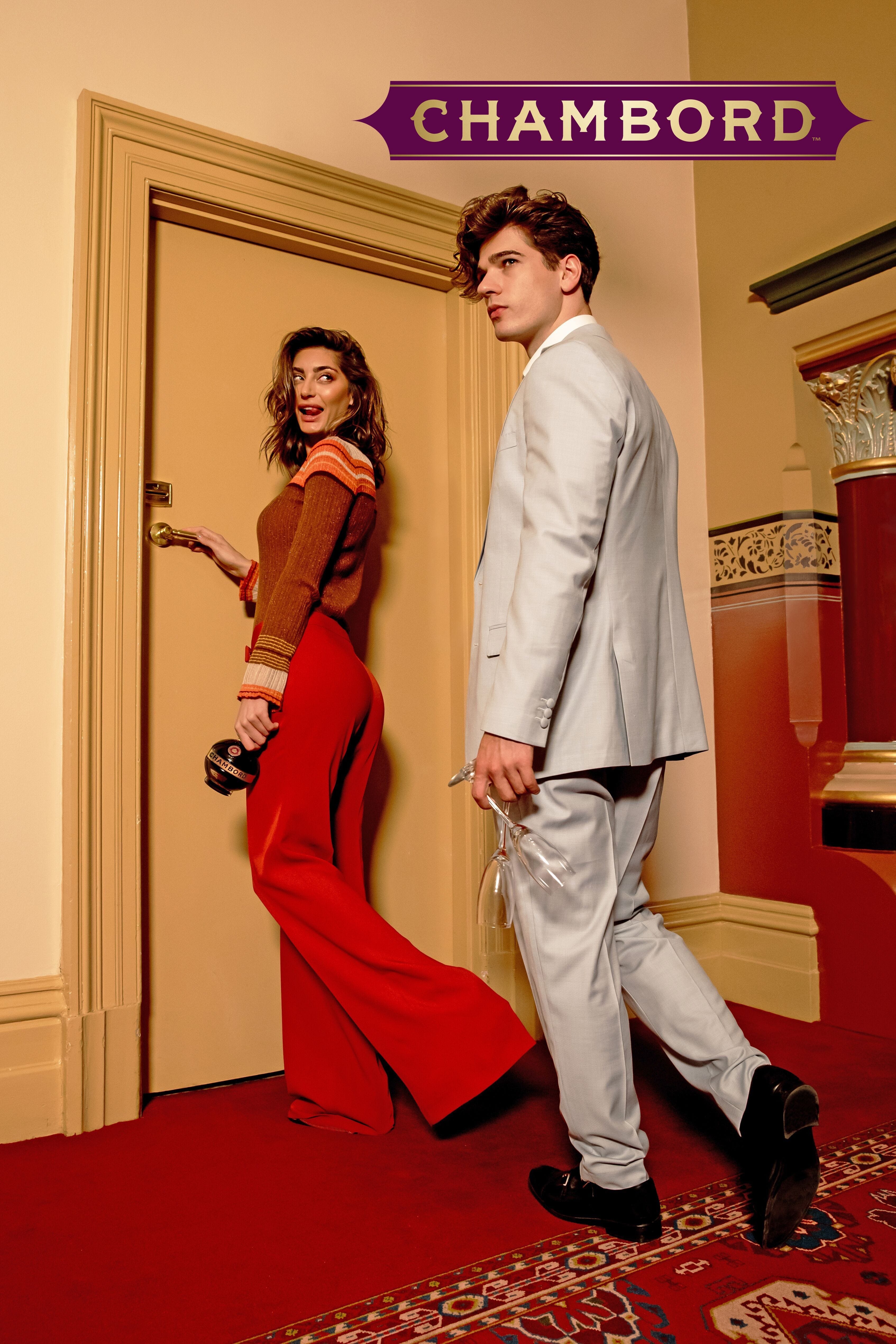 Stylish couple in a hotel corridor: a woman in red bell-bottoms walks toward a man in a silver suit, Chambord hotel signage above.