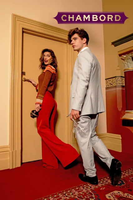 Stylish couple in a hotel corridor: a woman in red bell-bottoms walks toward a man in a silver suit, Chambord hotel signage above.