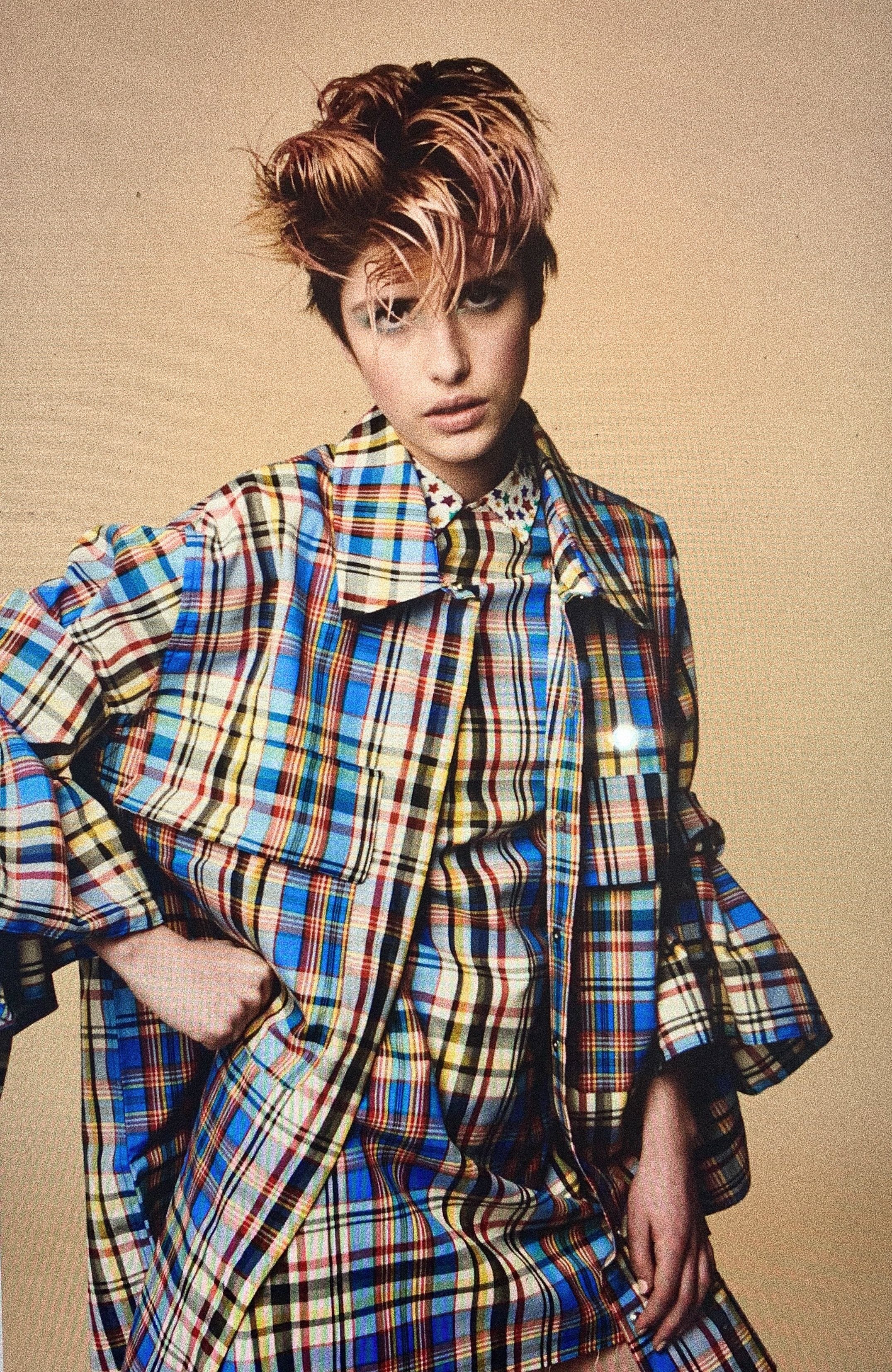 Fashion portrait of a young adult female model wearing a colorful plaid coat and dress in a studio with a neutral beige background.