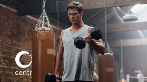 Adult male lifting dumbbells in a gym with punching bags visible in background, wearing a tank top and neutral expression.