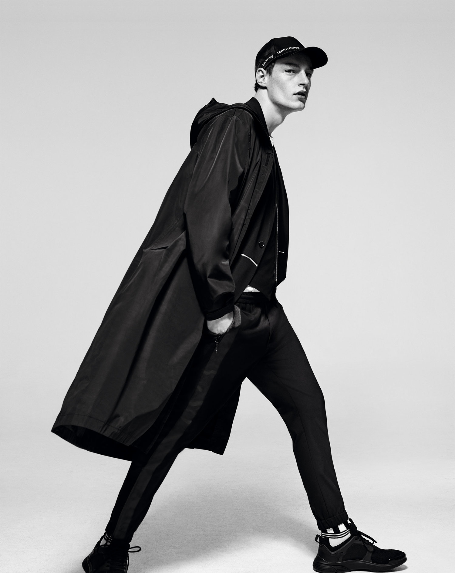 A male model in black sportswear strikes a walking pose against a neutral background, wearing a long coat, cap, pants, and sneakers.