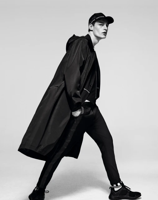 A male model in black sportswear strikes a walking pose against a neutral background, wearing a long coat, cap, pants, and sneakers.