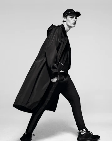 A male model in black sportswear strikes a walking pose against a neutral background, wearing a long coat, cap, pants, and sneakers.