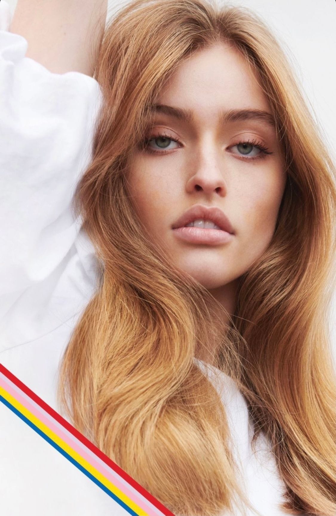 Close-up portrait of a young fair-skinned adult female with long strawberry blonde hair, wearing a white top with rainbow stripes.