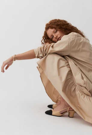 A red-haired woman in a neutral-toned dress and two-tone slingback heels poses in a photo studio against a white backdrop.