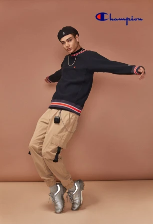 Young man strikes a pose in tan cargo pants, silver sneakers, and a navy sweatshirt, highlighted by a Champion logo and a neutral backdrop.