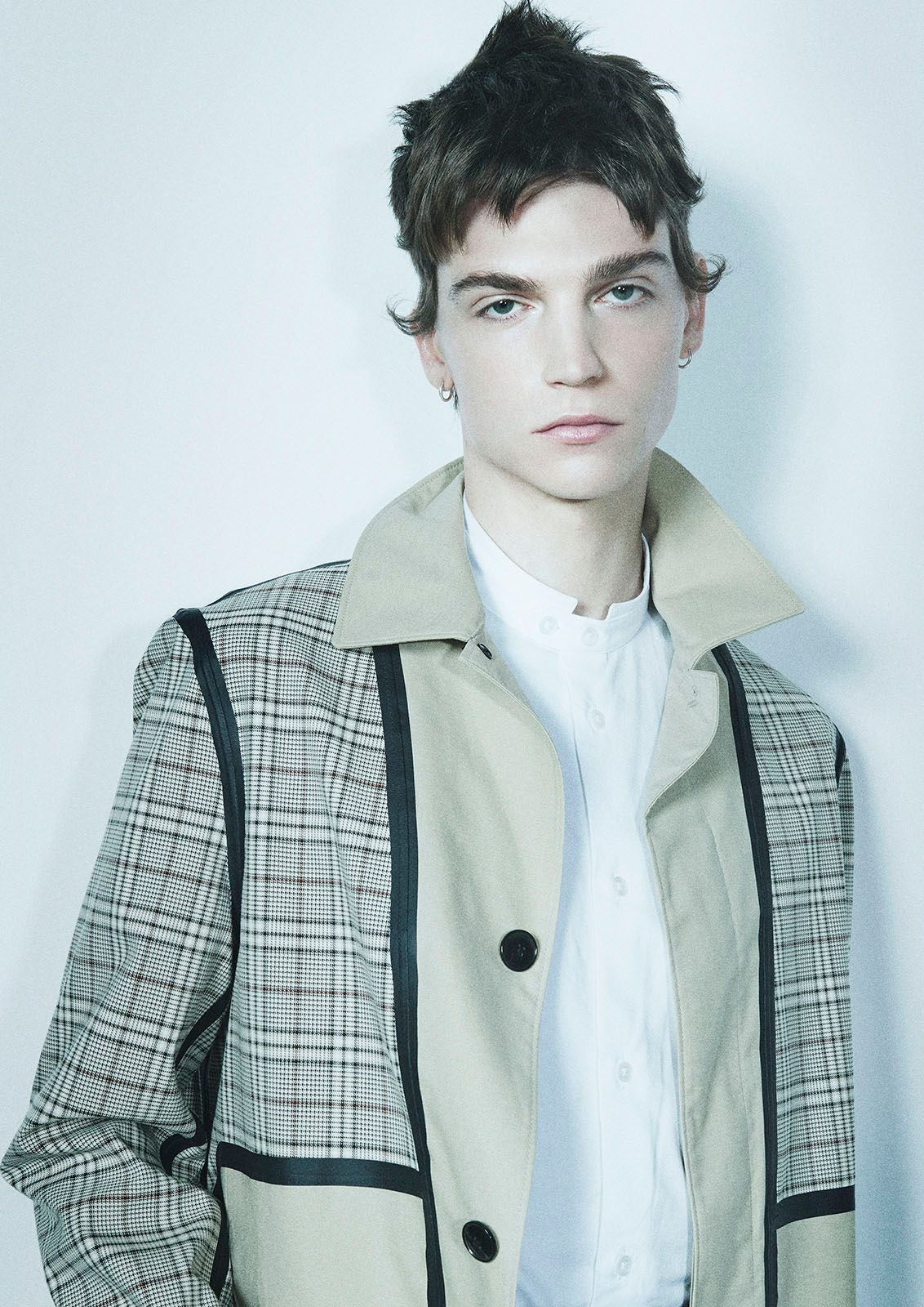 Portrait of a young adult male with a unique hairstyle, wearing a beige and plaid coat and a white collared shirt against a white backdrop.