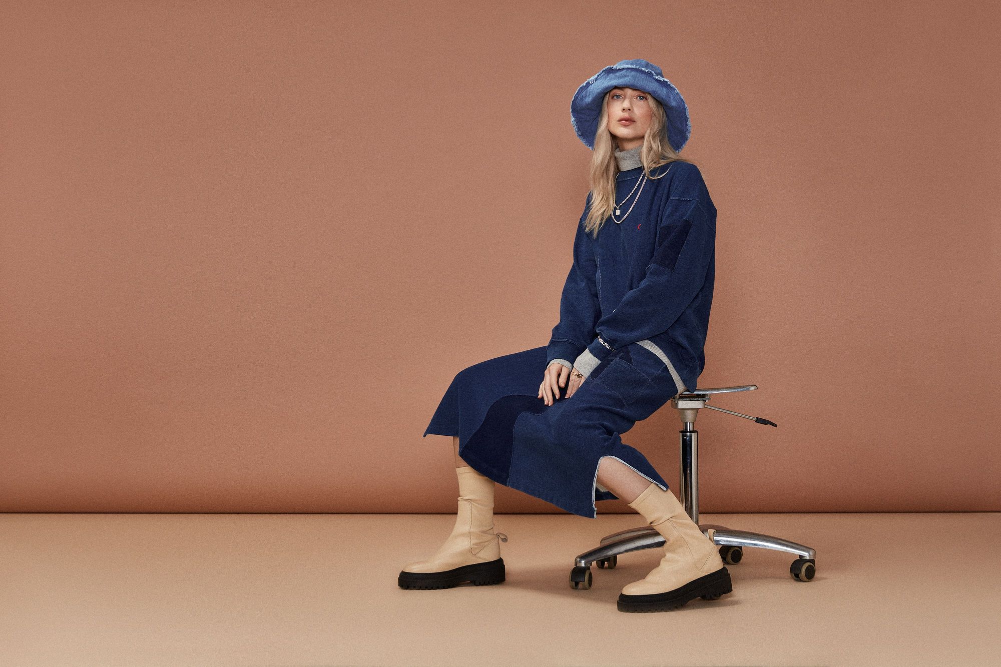 A fair-skinned woman is sitting on a rolling chair wearing a dark blue sweatshirt, skirt, bucket hat, and light-colored boots against a studio backdrop.