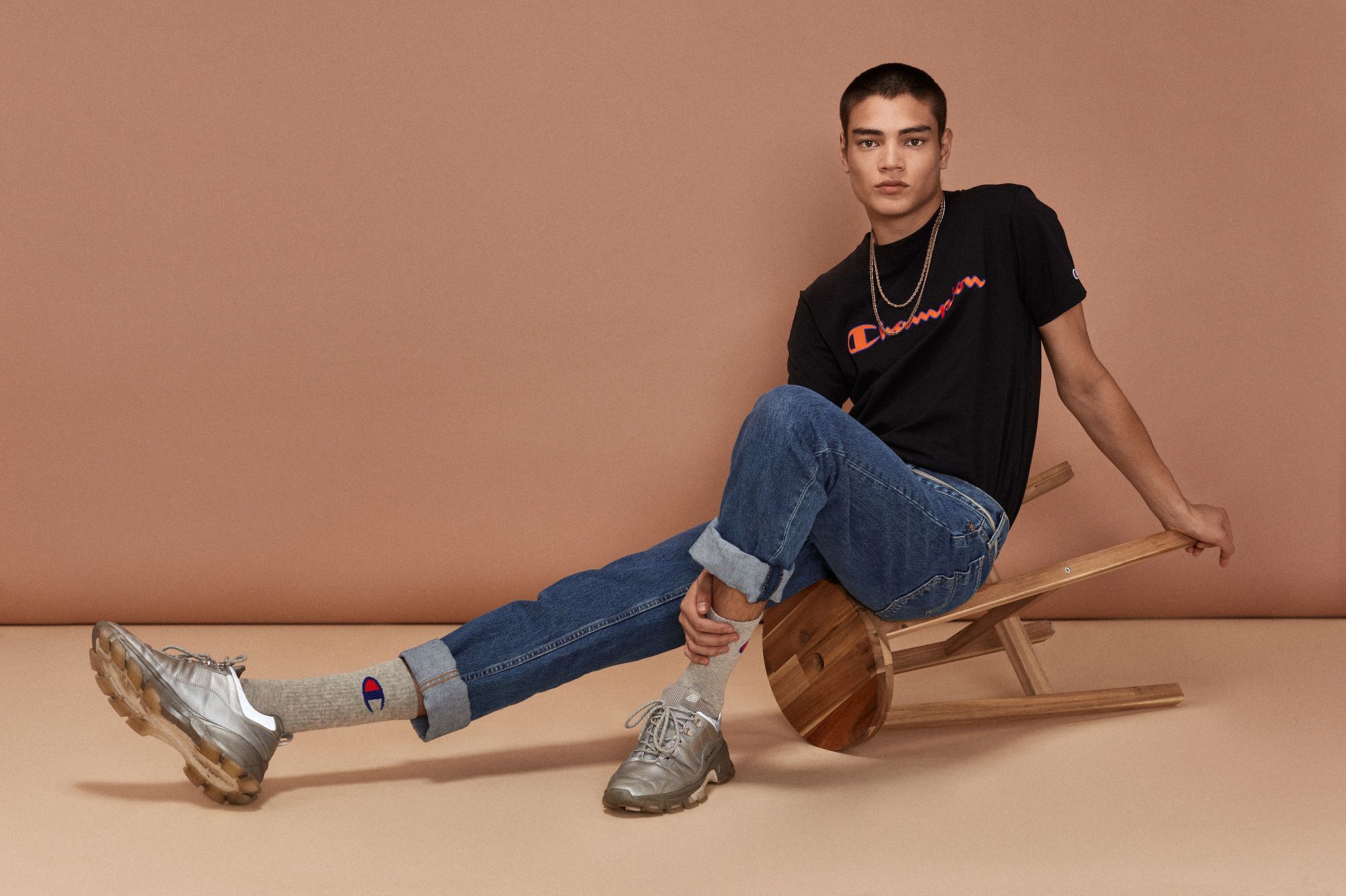 Man in Champion t-shirt and jeans sits on wooden stool indoors with a neutral background, showcasing casual fashion and style.