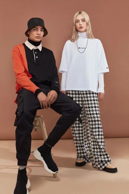 Two fashion models pose indoors against a brown backdrop, wearing Champion branded apparel with solid and patterned pants and bucket hats.