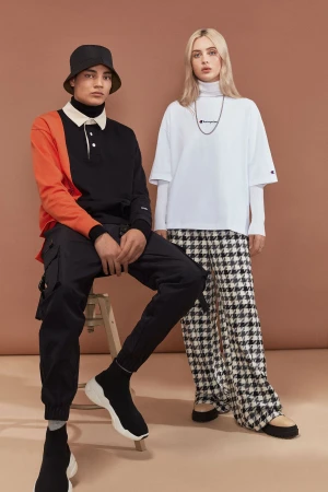 Two fashion models pose indoors against a brown backdrop, wearing Champion branded apparel with solid and patterned pants and bucket hats.