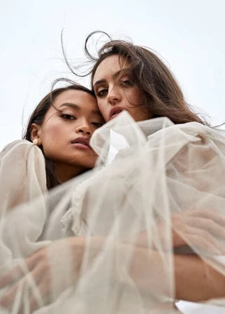 Low-angle portrait of two women wearing sheer off-white layered tulle garments against a pale overcast sky