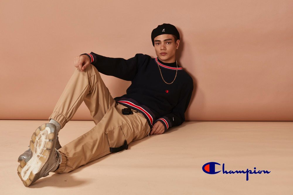 Young adult poses indoors wearing black Champion sweater, khaki pants, and black Kangol hat against a peach backdrop in studio shot.