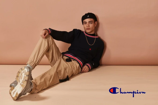 Young adult poses indoors wearing black Champion sweater, khaki pants, and black Kangol hat against a peach backdrop in studio shot.