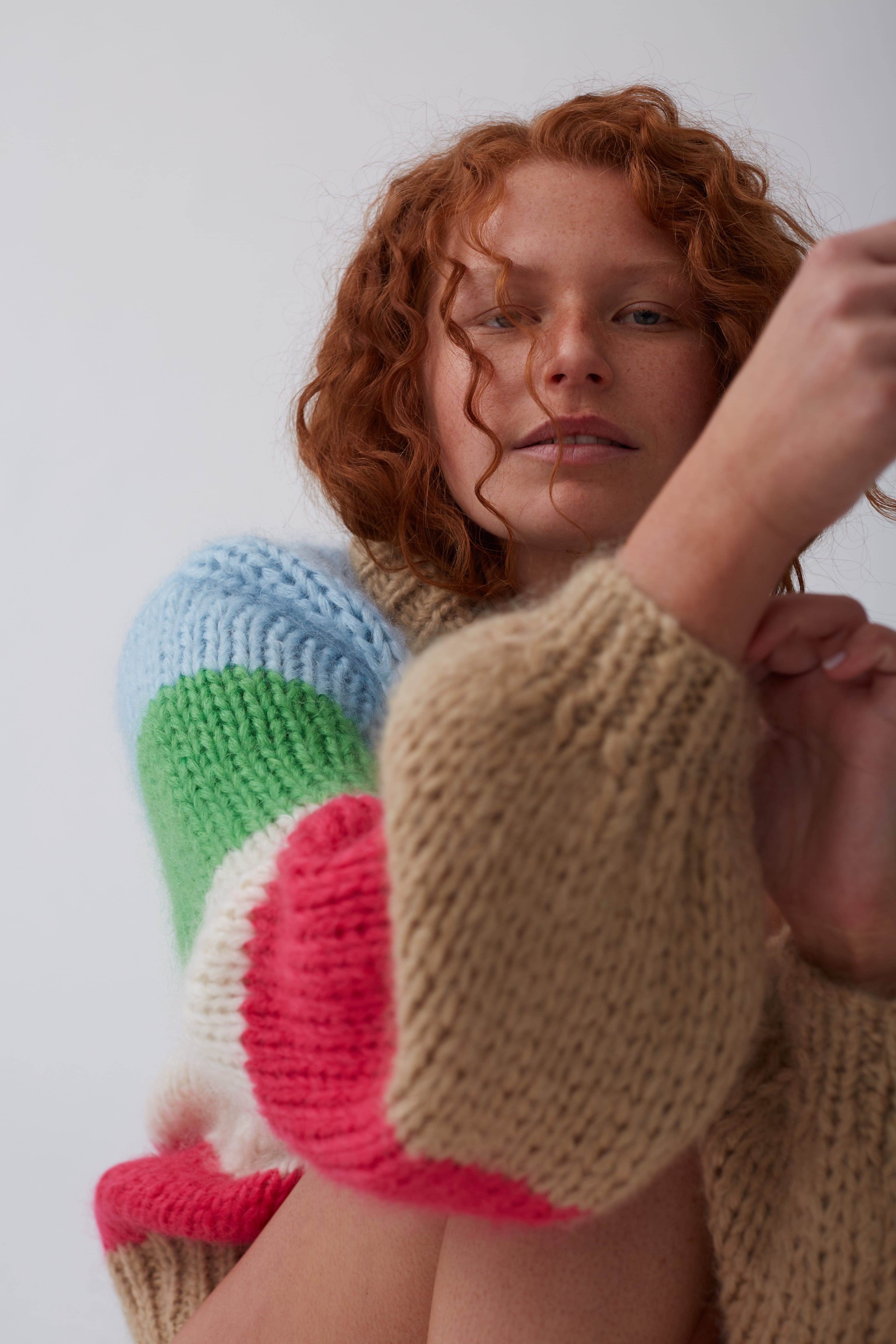 Portrait of a red-haired woman wearing a colour-blocked, chunky-knit sweater against a plain background in a medium studio shot.