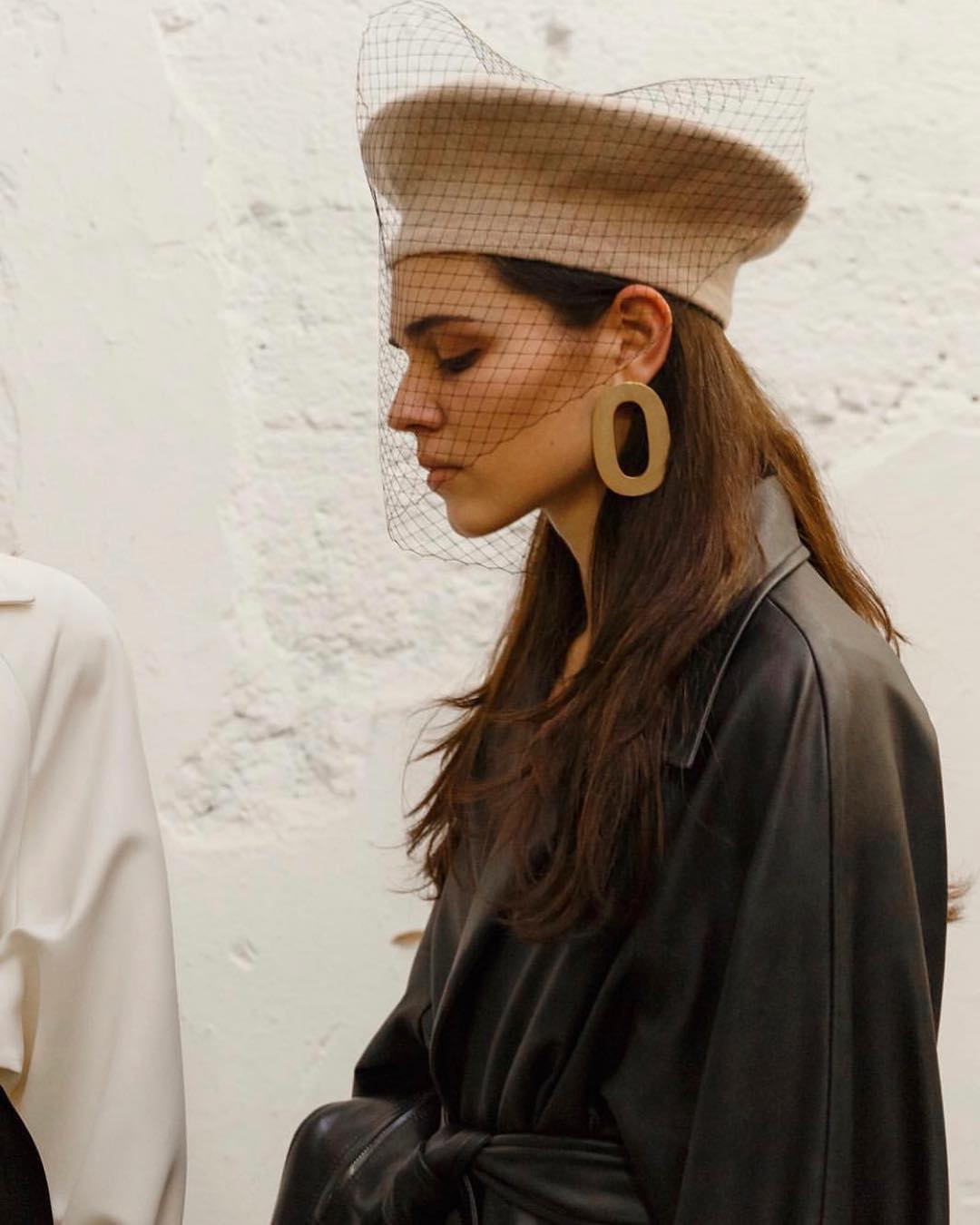 Profile shot of a woman wearing a beige beret with black netting and large earrings, posing in a black belted jacket indoors.