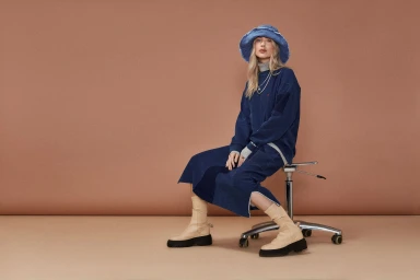 Model seated on a rolling stool wearing a blue denim skirt set, blue bucket hat, and light beige boots against a neutral backdrop.