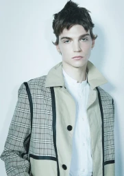 Close-up portrait of a young male model with dark messy hair wearing a tan plaid jacket and white shirt against a plain light wall.