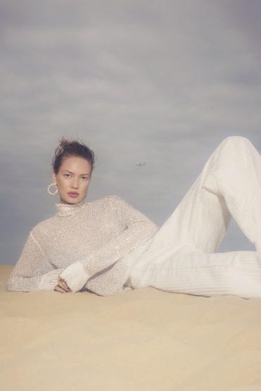 Woman in sheer top and white pants lying on sandy dune under blue sky, posing for outdoor fashion or beauty editorial photoshoot.