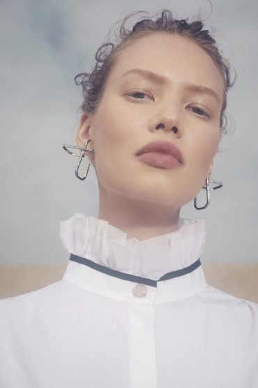 Close-up of a woman with curly hair, wearing statement earrings and a white, ruffled-collar blouse against a diffused blue and tan backdrop.