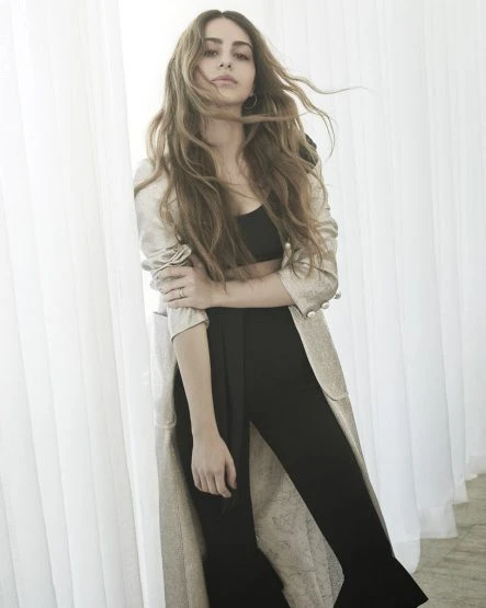Portrait of a young adult female with long brown hair wearing a beige coat, a black top, and black pants, posing indoors against a curtain.
