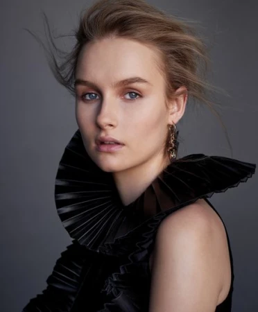 Portrait of a woman with blonde hair wearing a pleated black collar and drop earrings against a grey background.