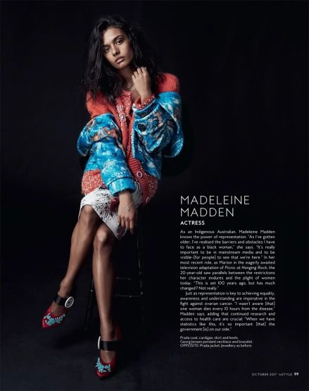Dark-haired woman in colorful patterned jacket sits on stool against dark background in a fashion editorial portrait shot.