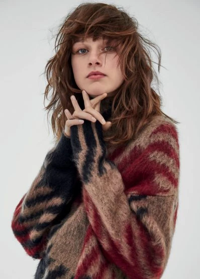 A young woman poses in a studio, wearing a multicolored patterned sweater. The focus is on her face and the texture of the knitwear.