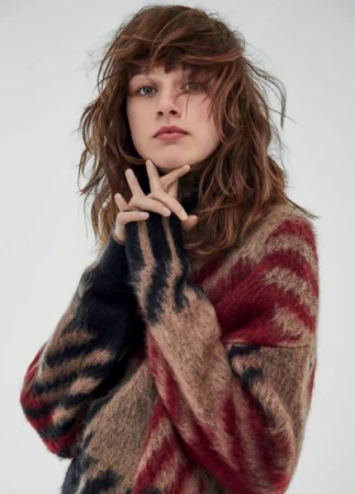 A young woman poses in a studio, wearing a multicolored patterned sweater. The focus is on her face and the texture of the knitwear.
