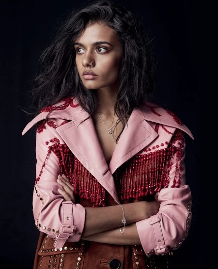 Portrait of a woman with dark hair wearing a pink leather coat with red fringe, set against a dark studio backdrop.
