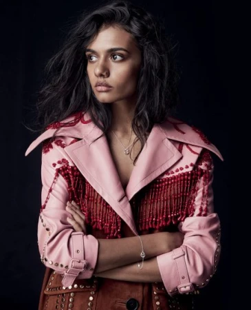 Portrait of a woman with dark hair wearing a pink leather coat with red fringe, set against a dark studio backdrop.