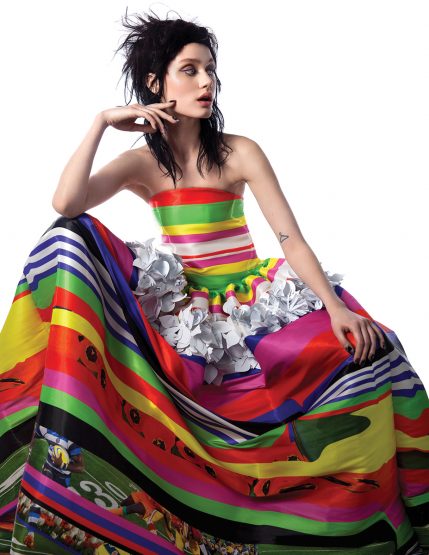 Woman in a colorful gown with rainbow stripes and white floral details, hand near her face, in a fashion style pose, against a white background.