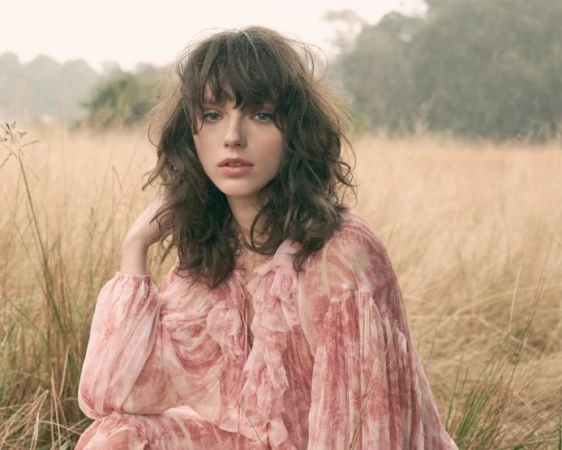 A woman with tousled dark hair and bangs wearing a pink patterned dress poses in a grassy meadow.