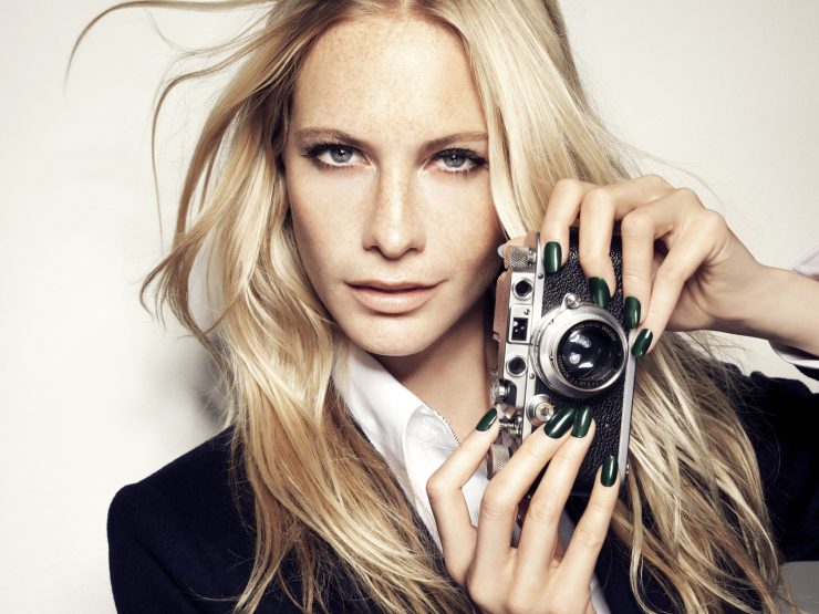 Blonde woman in a suit poses holding a vintage camera, showcasing green nail polish against a plain white background in a studio setting.